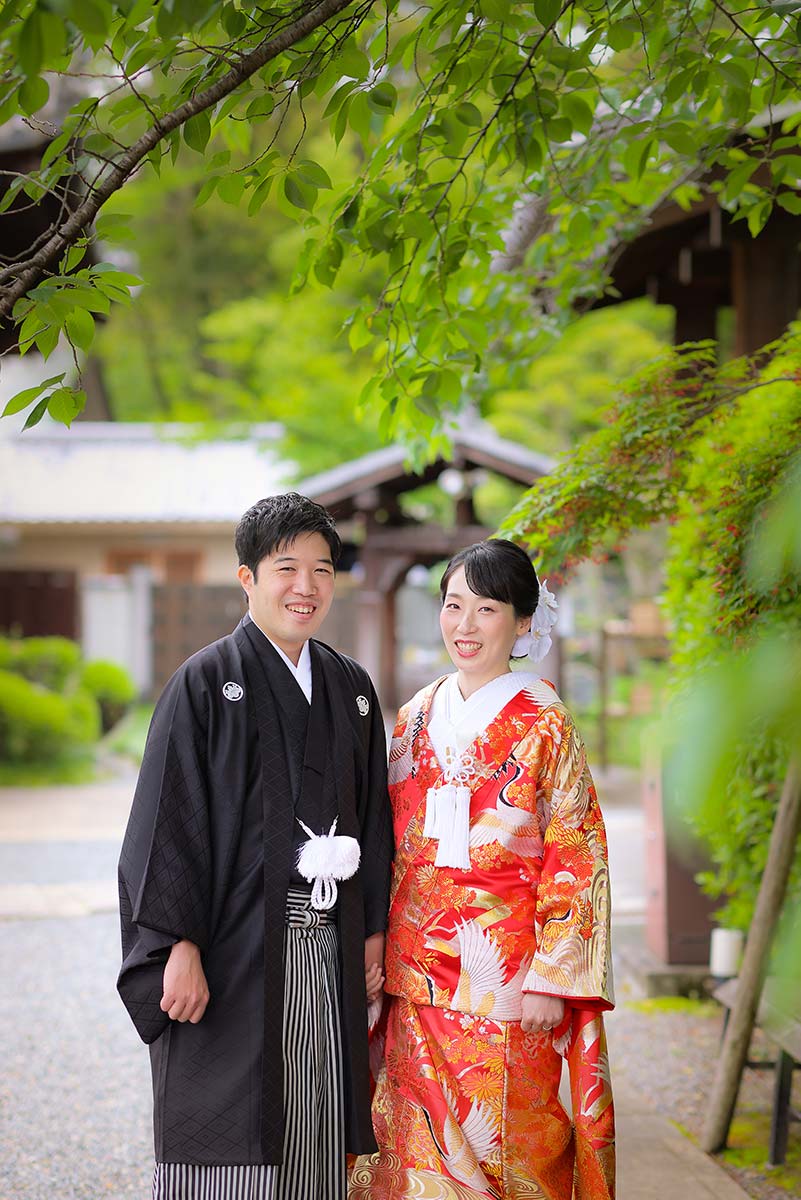 桜の木の緑を背景に笑顔の色打掛羽織袴姿の新郎新婦様